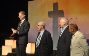 four men stand on stage addressing an audience